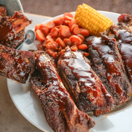 A plate of glazed ribs with carrots and corn on the cob, served on a rustic table.