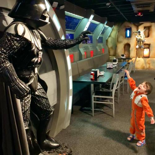A child in an orange jumpsuit looks up in awe at a life-sized Darth Vader figure in a themed restaurant.
