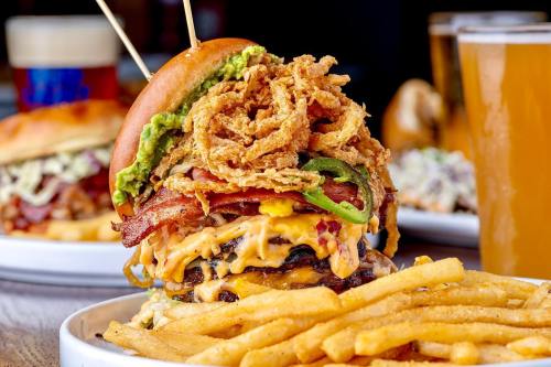 A towering burger with layers of cheese, bacon, jalapeños, and crispy onions, served with a side of fries.