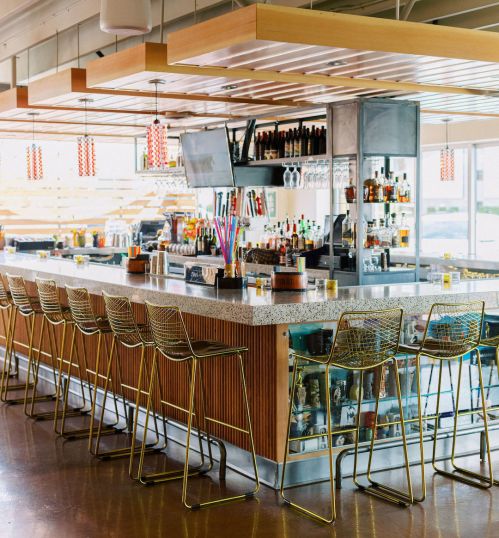 A modern bar with a long countertop, high stools, and shelves stocked with drinks, featuring bright pendant lights above.