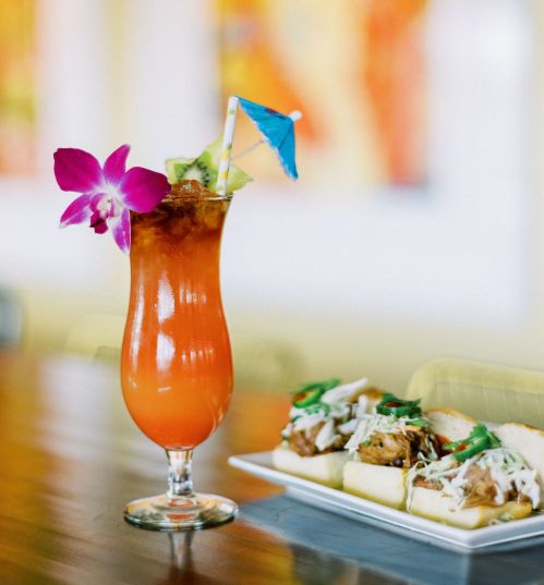 A colorful tropical drink garnished with an orchid and a small umbrella, alongside a plate of savory food.