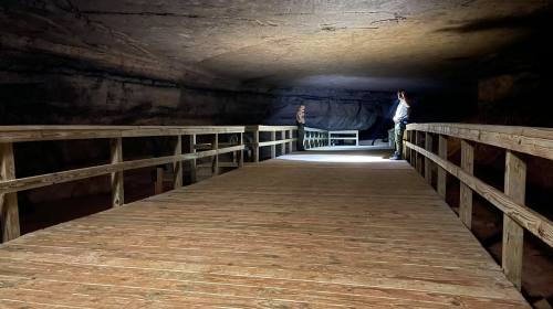 Two people stand on a wooden walkway inside a dimly lit cave, with light illuminating the path ahead.