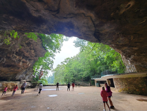 A group of people exploring a large cave opening surrounded by lush green trees and a sandy floor.
