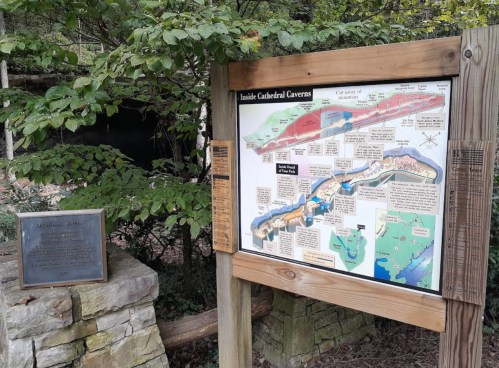 Map of Cathedral Caverns displayed on a wooden sign, surrounded by greenery and a plaque nearby.