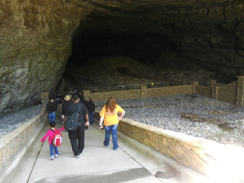 Visitors walking towards a large cave entrance, surrounded by rocky terrain and stone walls.