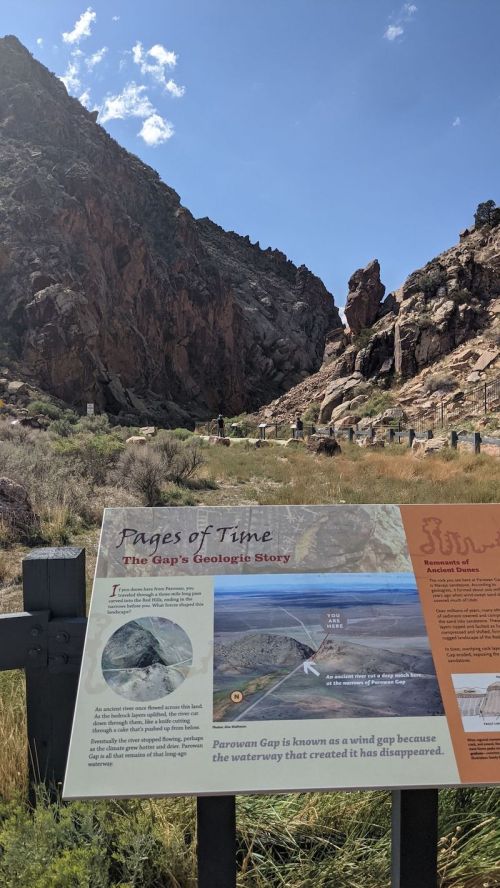 Sign about Parowan Gap's geology, with rocky cliffs and a grassy area in the background under a clear blue sky.