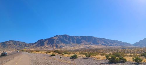 A wide view of a desert landscape with mountains in the background and a clear blue sky. A road runs alongside the scene.