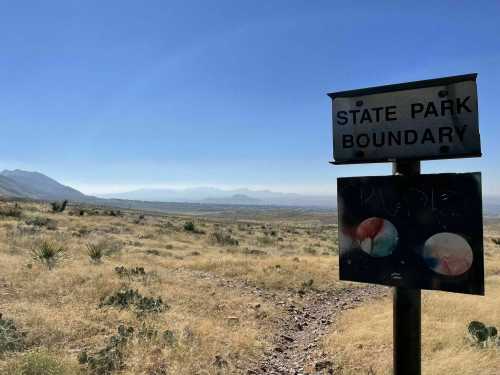 A weathered sign marks the boundary of a state park, with a vast, dry landscape and distant mountains under a clear sky.