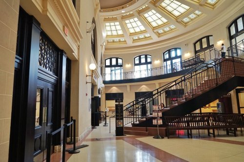 Interior of a spacious, well-lit building featuring a grand staircase, large windows, and decorative ceiling.