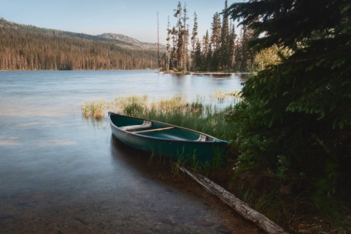 A green canoe rests on the shore of a tranquil lake, surrounded by trees and mountains in the background.