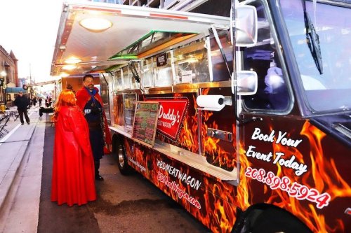 A food truck named "Daddy's" with a fiery design, featuring a vendor in a superhero costume and a customer in a red cape.