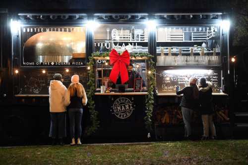 A food truck decorated for the holidays, with a large bow, serving pizza to customers in a festive outdoor setting.