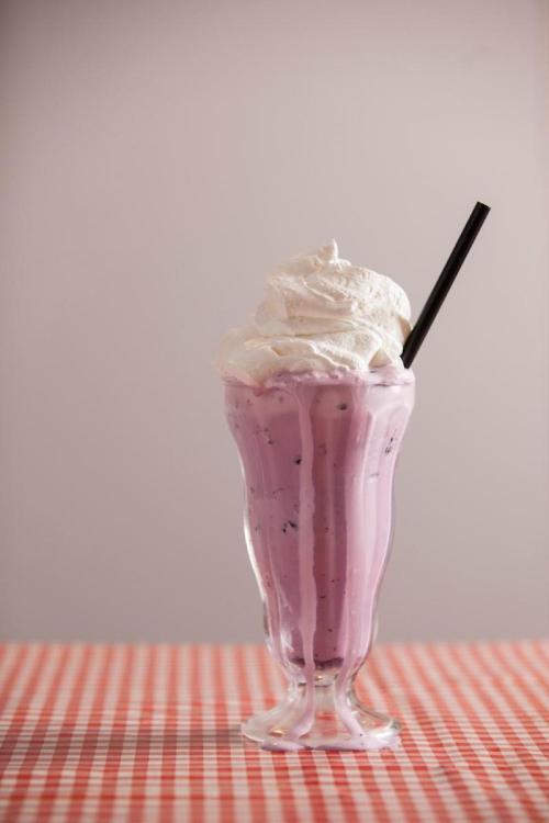 A tall glass of purple milkshake topped with whipped cream and a black straw, set on a checkered tablecloth.