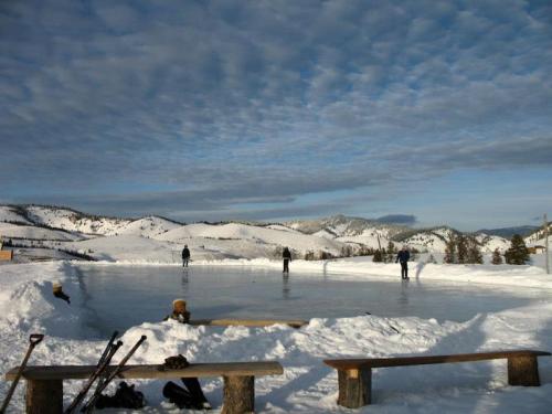 A snowy landscape with an outdoor ice rink and people skating, surrounded by mountains and a cloudy sky.