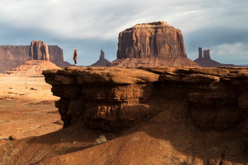 A person stands on a rocky ledge overlooking a vast desert landscape with towering red rock formations.