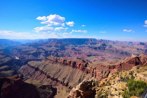 A panoramic view of the Grand Canyon, showcasing layered rock formations and a clear blue sky with scattered clouds.