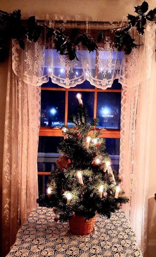 A small Christmas tree with candles sits on a lace tablecloth, illuminated by soft light from a window.