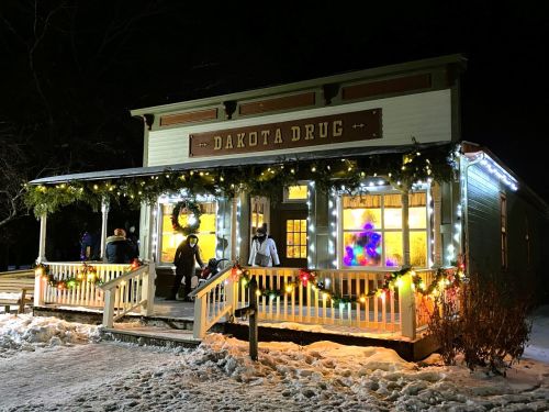 A festive storefront of Dakota Drug, adorned with holiday lights and decorations, set against a snowy night backdrop.