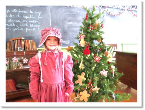 A young girl in a pink dress and bonnet stands beside a decorated Christmas tree in a classroom setting.