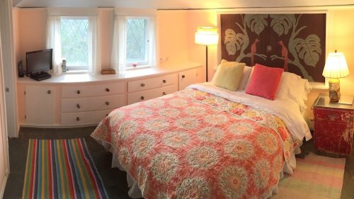 Cozy bedroom with a colorful quilt, two windows, a TV, and a lamp, featuring a vibrant rug and decorative wall art.