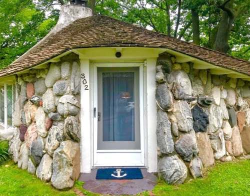 A unique stone cottage with a rounded roof and a front door, featuring an anchor doormat and surrounded by greenery.