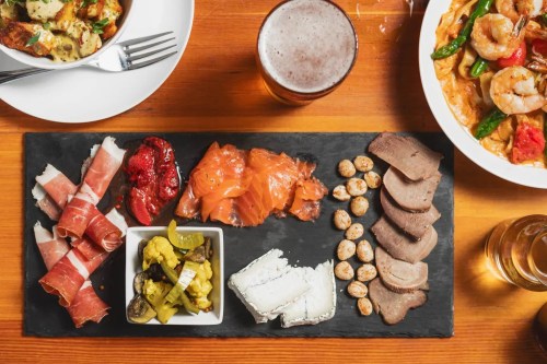 A wooden table with a charcuterie board featuring meats, cheeses, pickles, and nuts, alongside a beer and a seafood dish.