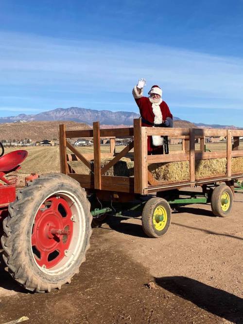 Santa Claus waves from a wooden wagon filled with hay, parked next to a red tractor against a clear blue sky.