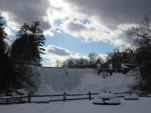 A snowy landscape featuring a frozen waterfall under a cloudy sky, with trees and a wooden fence in the foreground.
