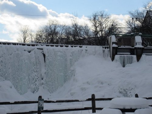 Frozen waterfall cascading over a dam, surrounded by snow and trees under a partly cloudy sky.