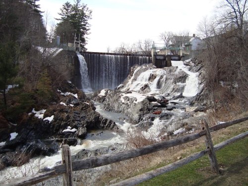 A scenic view of a waterfall cascading over rocks, with a dam and trees in the background on a clear day.