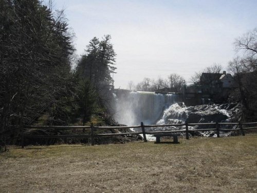A scenic view of a waterfall surrounded by trees and a wooden fence, with a clear sky in the background.
