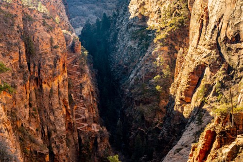 A narrow canyon with steep, rocky walls and winding trails visible along the slopes, illuminated by soft sunlight.