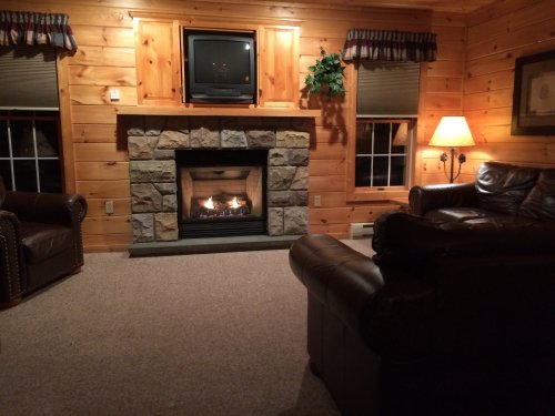 Cozy living room with a stone fireplace, wooden walls, a TV, and comfortable leather furniture. Warm and inviting atmosphere.