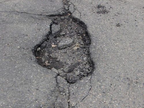 A large pothole in a paved road, revealing loose gravel and cracked asphalt around the edges.
