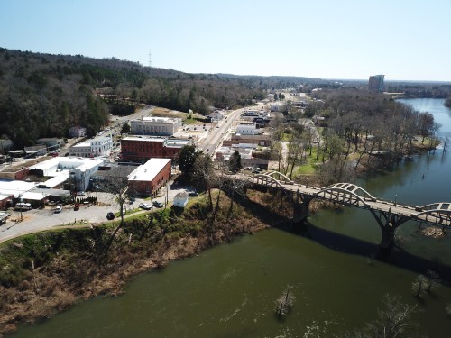 Aerial view of a small town by a river, featuring buildings, a bridge, and tree-covered hills in the background.