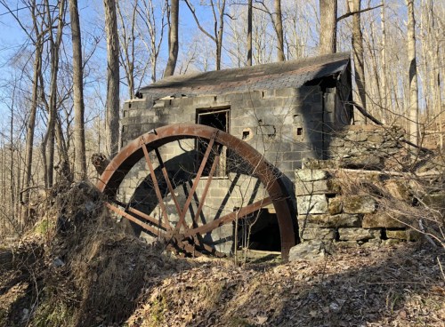 Abandoned structure with a large waterwheel, surrounded by trees and underbrush in a wooded area.