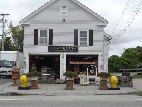 A white antique shop with black shutters, featuring outdoor decor and planters, set along a road with greenery in the background.