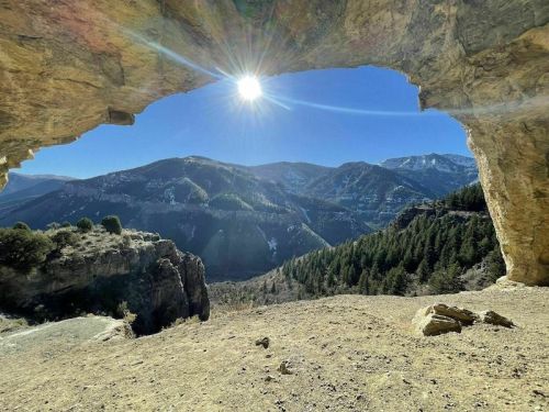 A sunlit view from a rocky arch, overlooking a valley with mountains and trees under a clear blue sky.