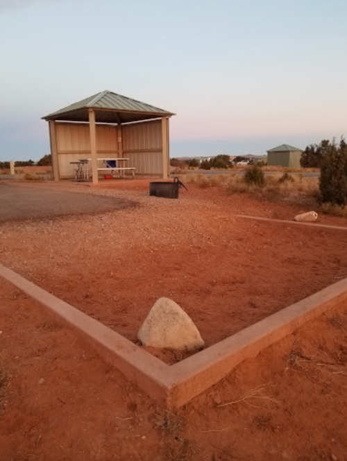 A small shelter with a metal roof in a desert landscape at sunset, surrounded by red earth and sparse vegetation.