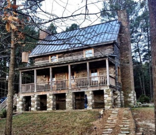 A rustic two-story cabin with a metal roof, stone foundation, and wooden porch, surrounded by trees.