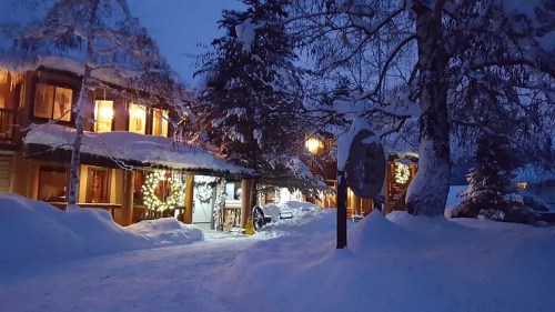 A snowy scene at dusk featuring cozy cabins adorned with lights and surrounded by trees.