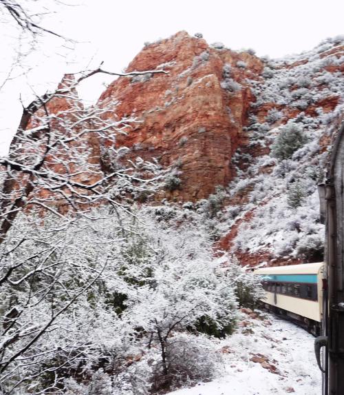 A train travels through a snowy landscape, surrounded by rocky cliffs and evergreen trees.