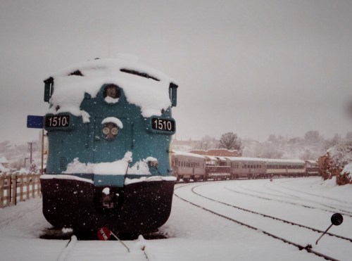 A snow-covered train engine stands at a station, with another train visible in the background amidst falling snow.