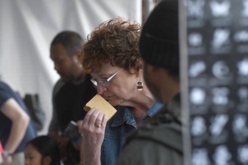 A woman with curly hair and glasses is tasting a food item at a crowded event, with people in the background.