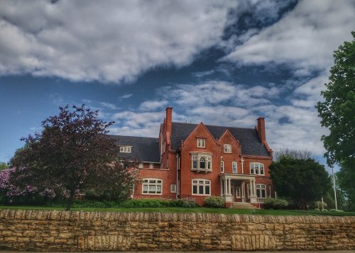 A large red brick house with multiple windows, surrounded by greenery and under a partly cloudy sky.
