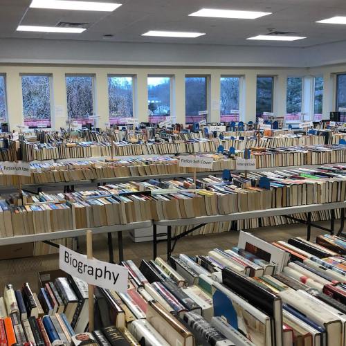 A spacious room filled with tables stacked high with books, organized by genre, including a "Biography" section.