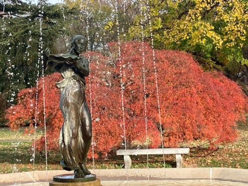 A bronze statue of a woman stands in a fountain, with vibrant red foliage in the background.