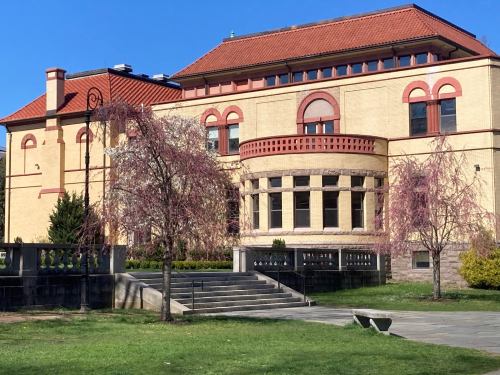 A historic building with a red-tiled roof, large windows, and blooming trees in a green park setting.
