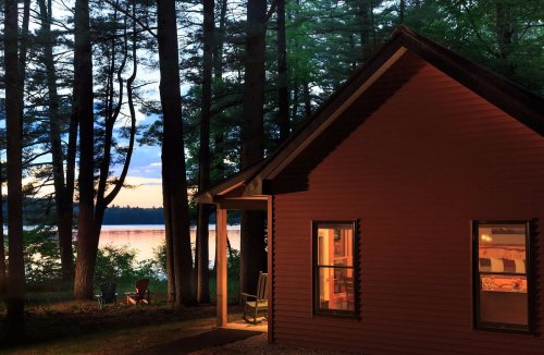 A cozy cabin by a lake at dusk, surrounded by trees, with warm light glowing from the windows.