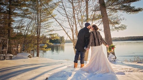 A couple kisses in a snowy landscape by a lake, surrounded by trees, celebrating their wedding.